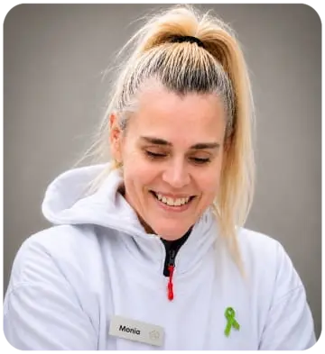 Monia Berg sitzt leicht nach vorne gebeugt. Sie hat helle, zu einem Pferdeschwanz gebundene Haare und trägt einen weissen Hoodie und die "grüne Schleife". 